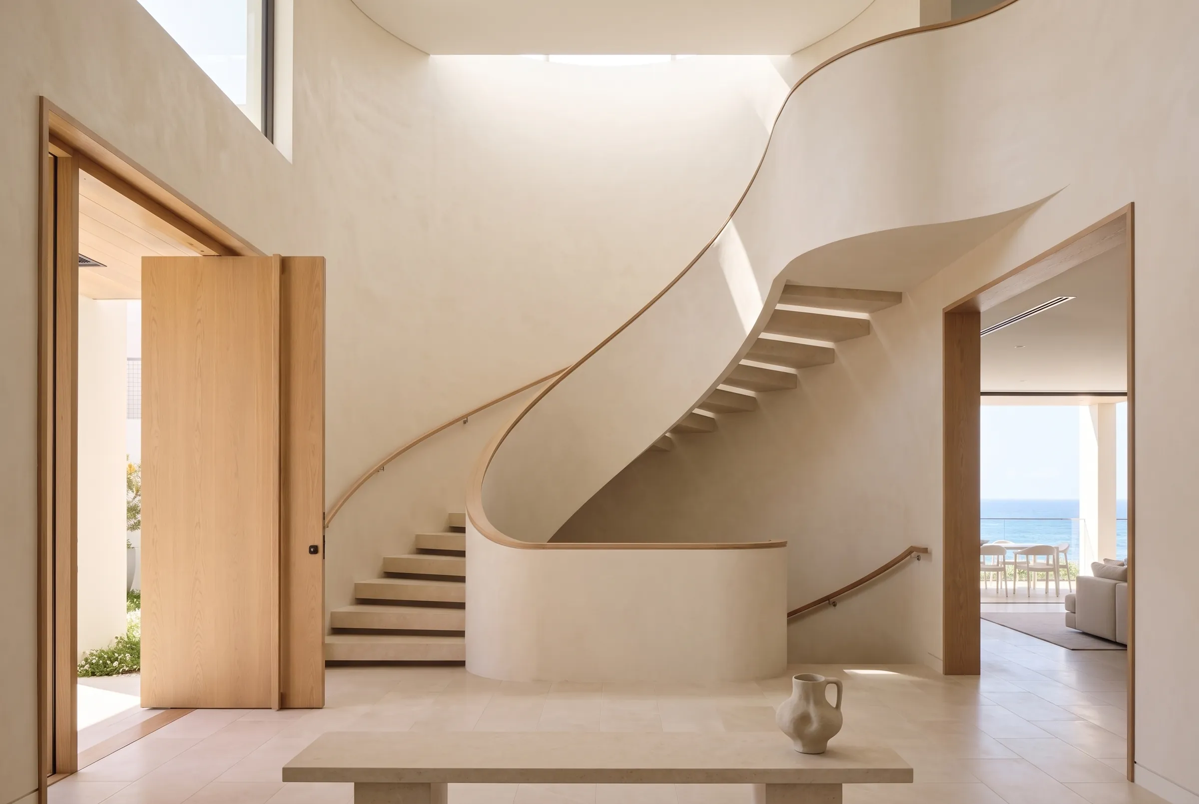 Interior design visualisation of a sculptural curved staircase in warm lime render with limestone treads and an oak handrail, beside an oak pivot front door